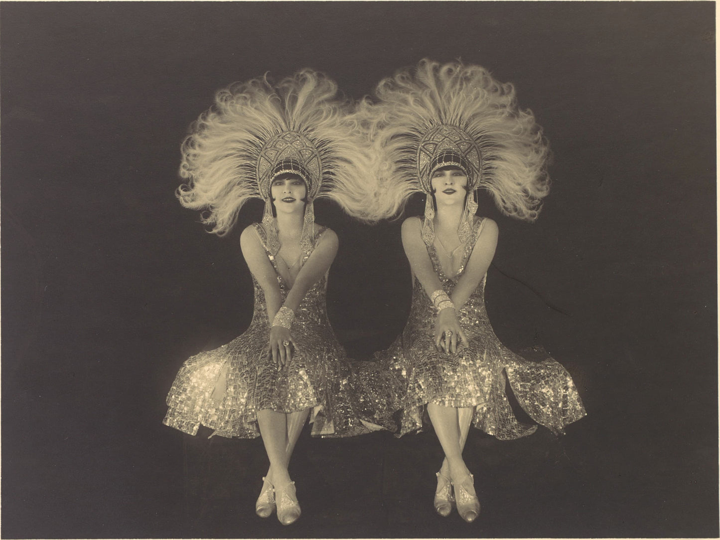 Two women in sparkling dresses and large feathered headpieces on a dark background