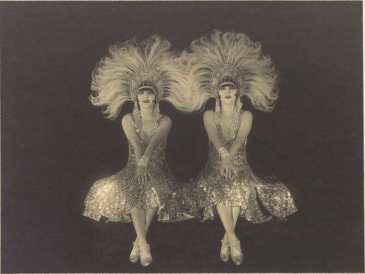 Two women in sparkling dresses and large feathered headpieces on a dark background