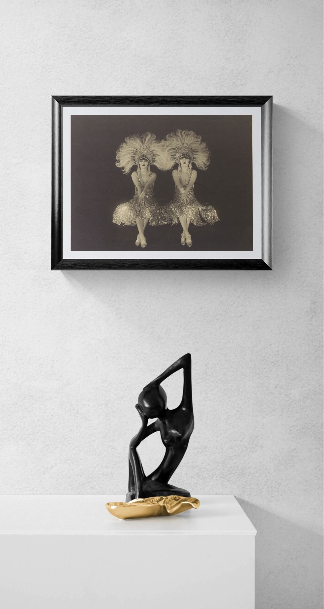 Framed artwork of two dancers on a white wall with a black sculpture below.