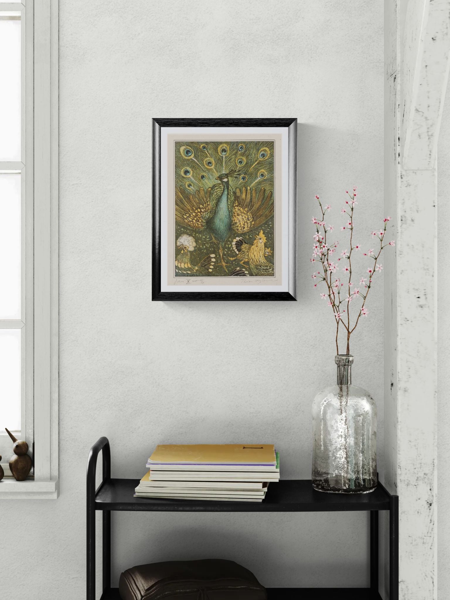Framed peacock artwork on a wall above a black shelf with books and a vase.