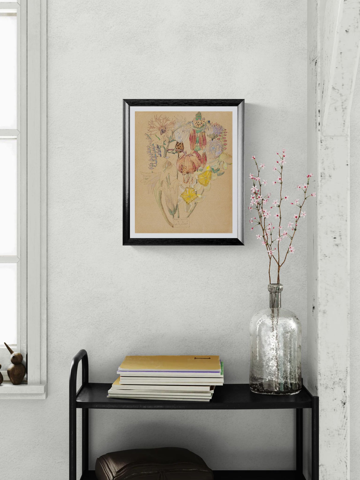 Framed artwork on a wall above a black shelf with books and a vase.