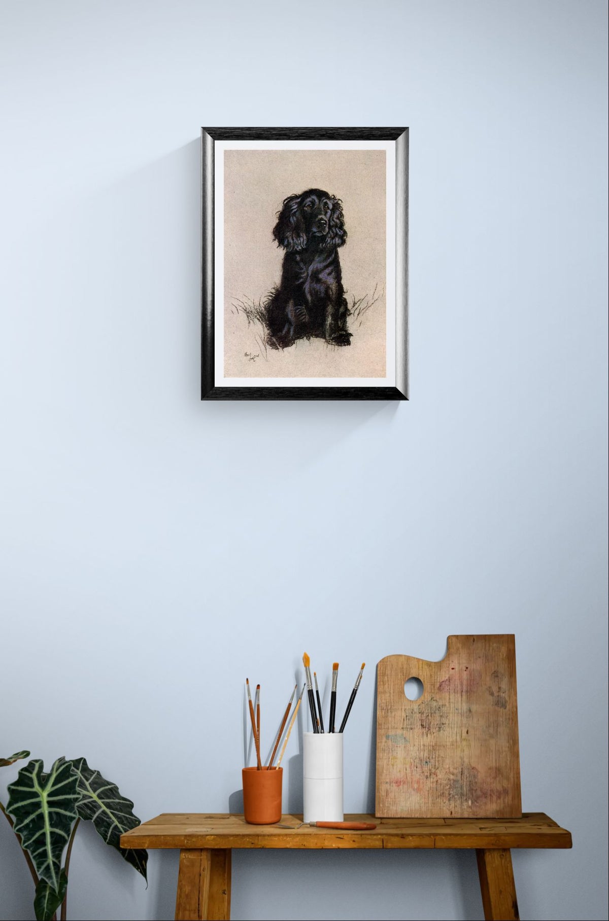 Framed artwork of a black dog on a light blue wall above a wooden table with art supplies.