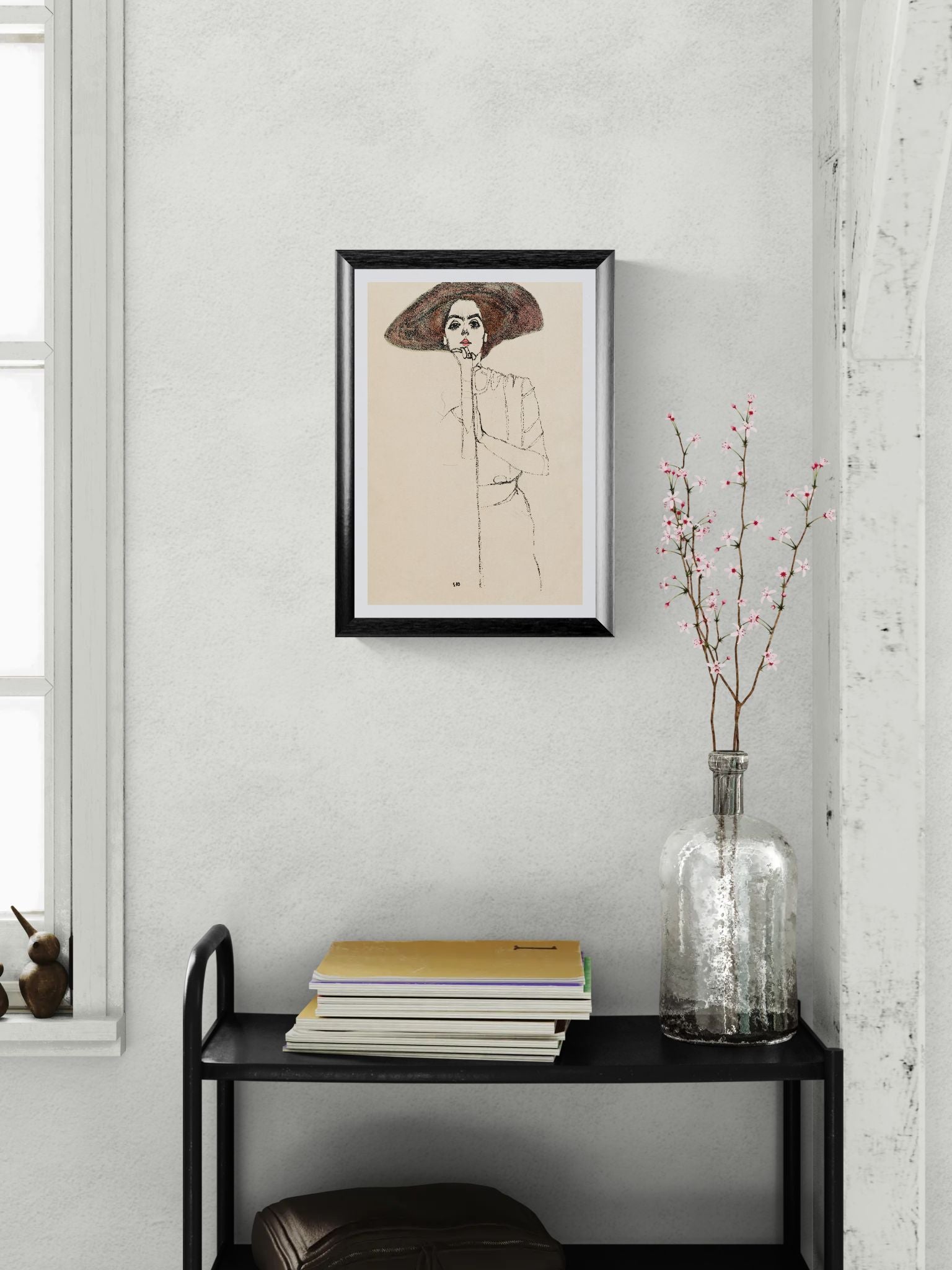 Framed artwork on a wall above a black shelf with books and a vase.