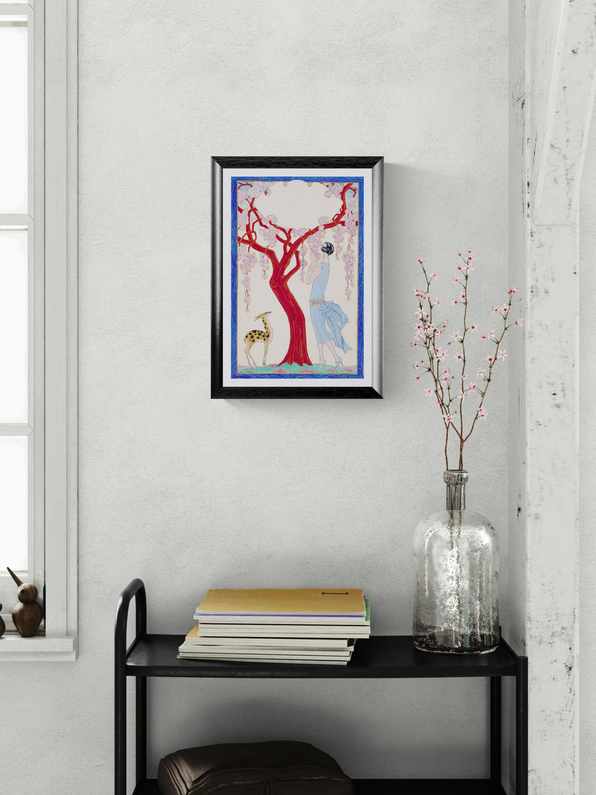 Framed artwork on a wall above a black shelf with books and a vase.