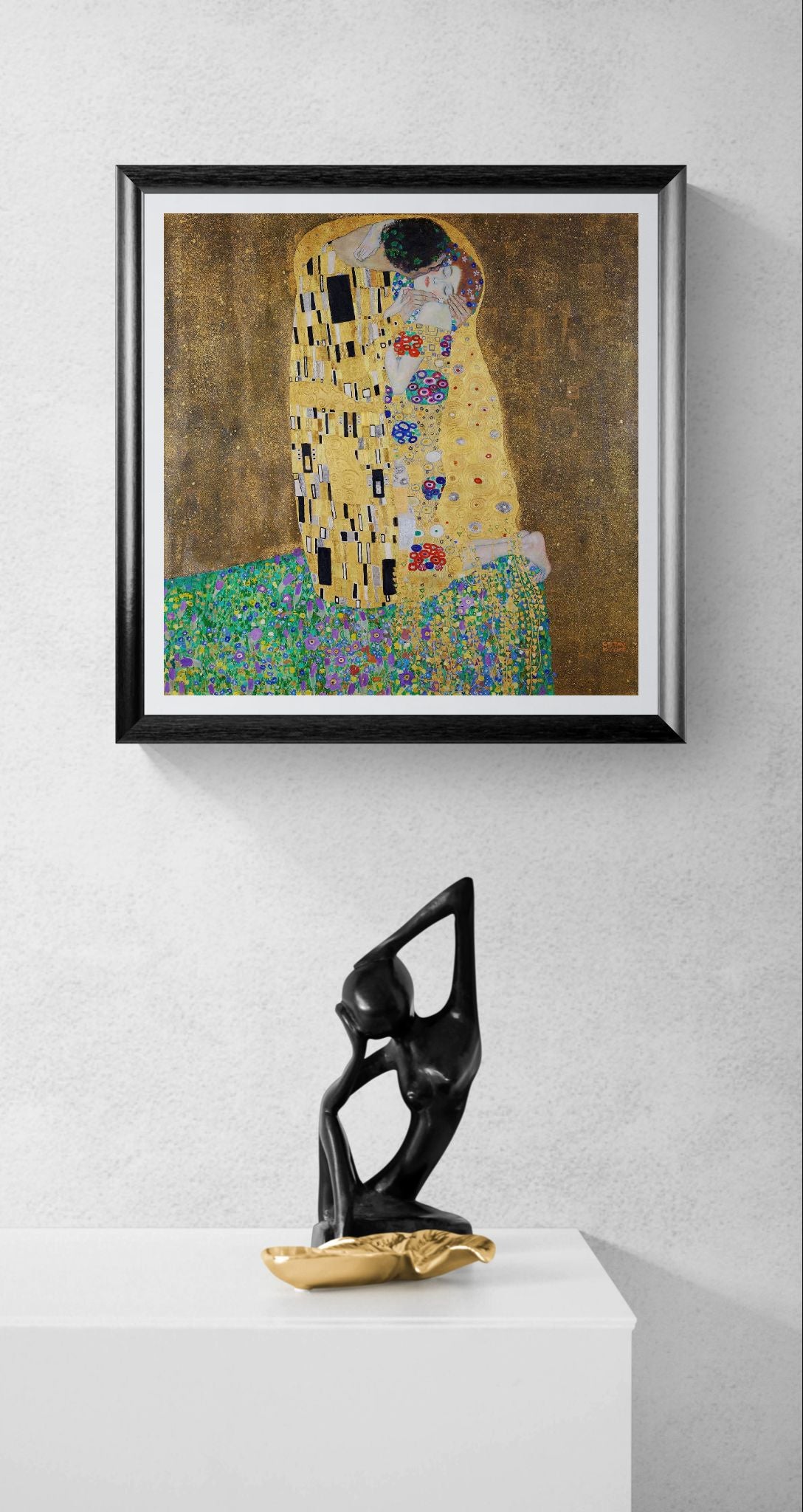 Framed artwork resembling 'The Kiss' by Gustav Klimt with a black and gold sculpture below on a white surface.