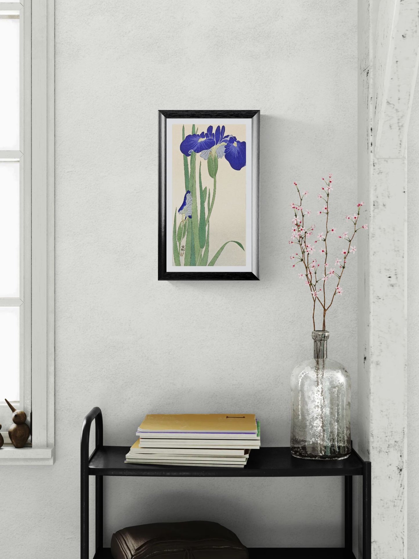 Framed artwork of flowers on a wall above a black shelf with books and a vase.