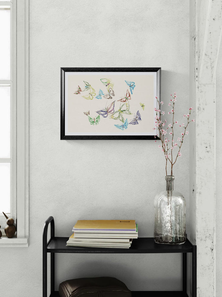 Framed artwork of colorful butterflies on a white wall above a black shelf with books and a vase.