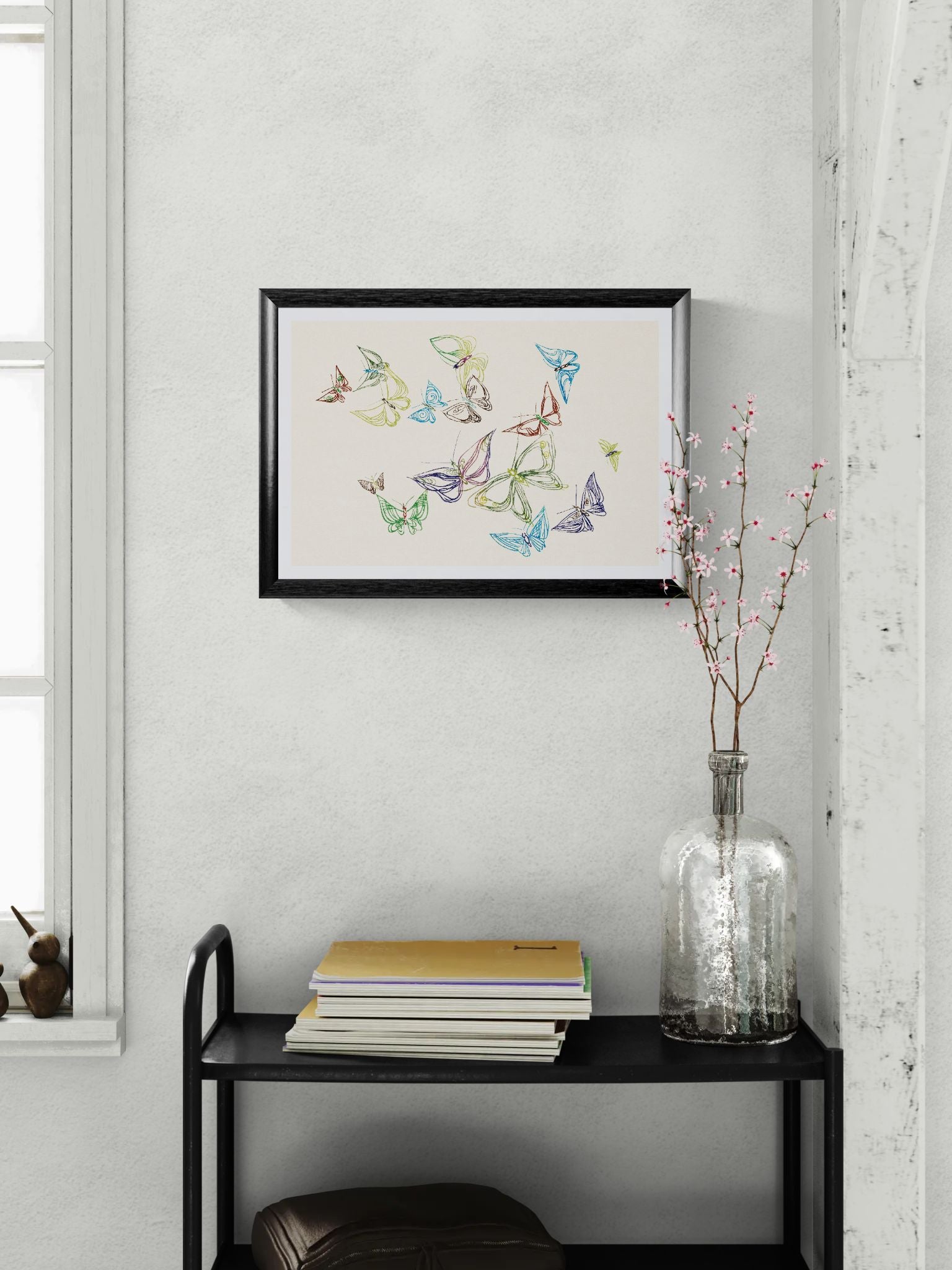 Framed artwork of colorful butterflies on a white wall above a black shelf with books and a vase.