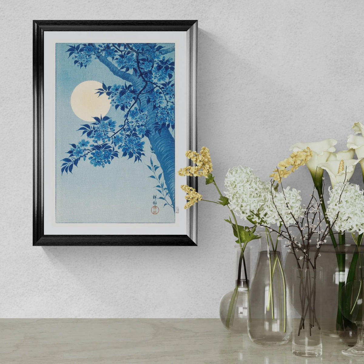 Framed artwork with blue floral design and moon on a wall above glass vases with flowers.