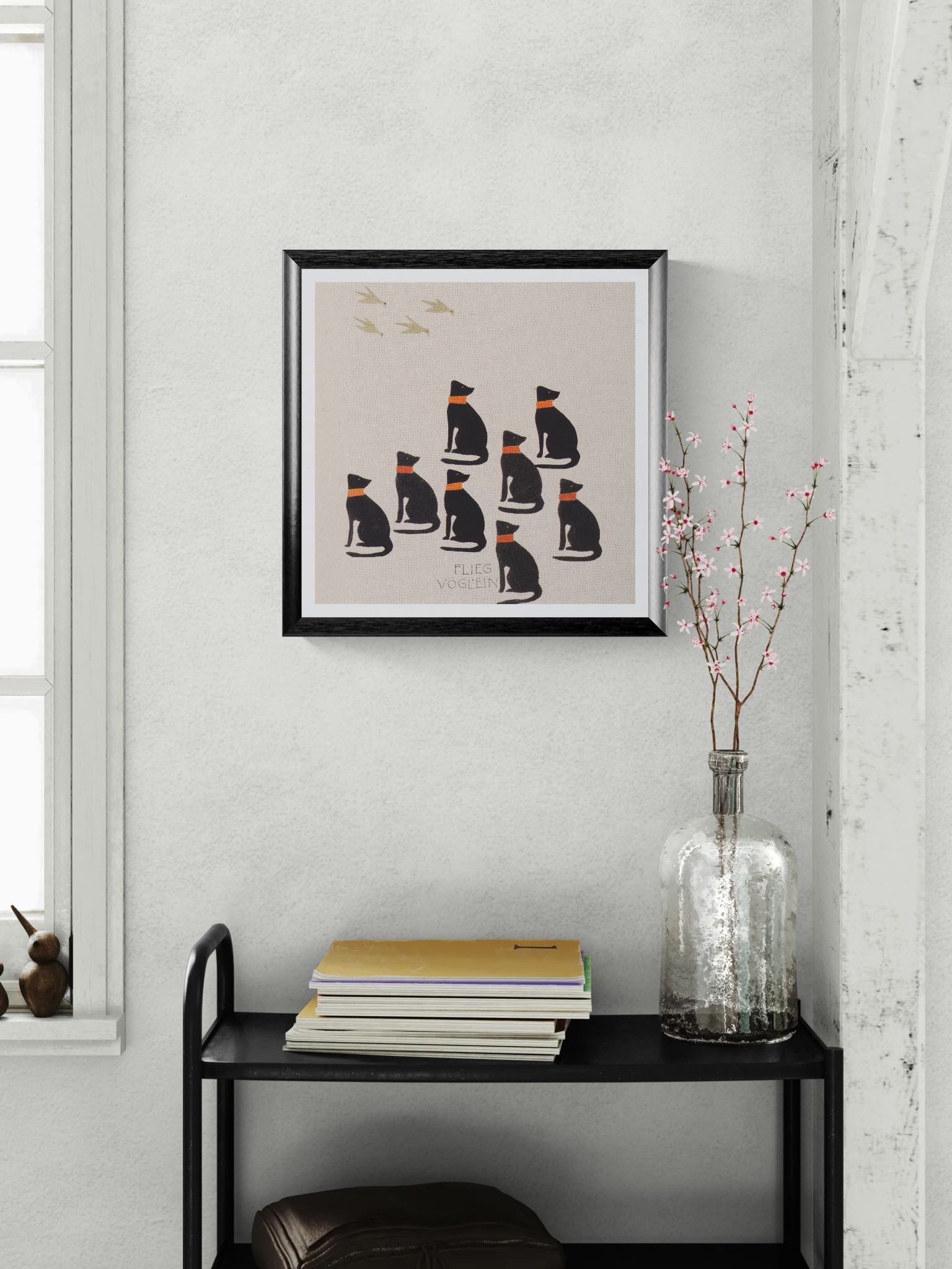 Framed artwork of cats on a wall above a black shelf with books and a vase.
