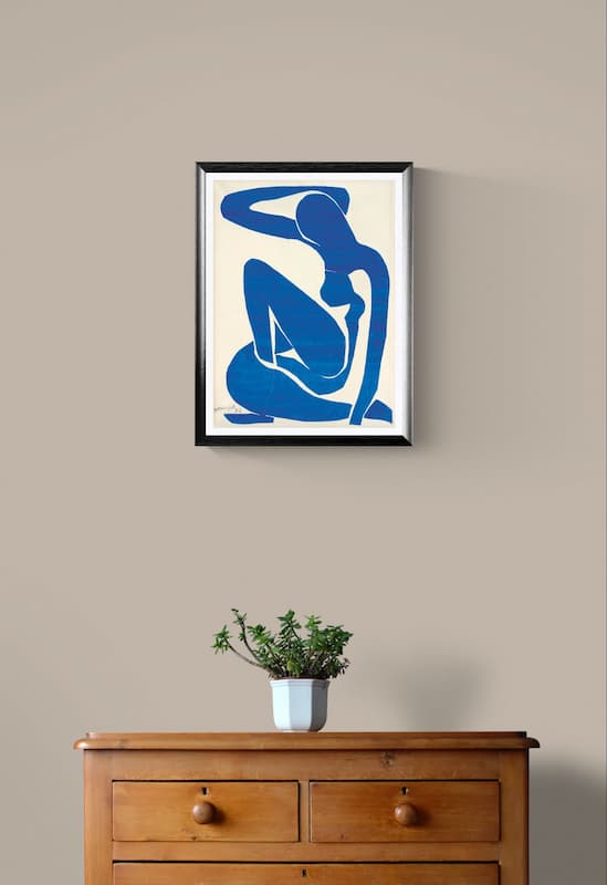 A framed abstract art print featuring a blue silhouette of a seated figure on a cream background, hanging on a beige wall above a wooden cabinet with a small potted plant.