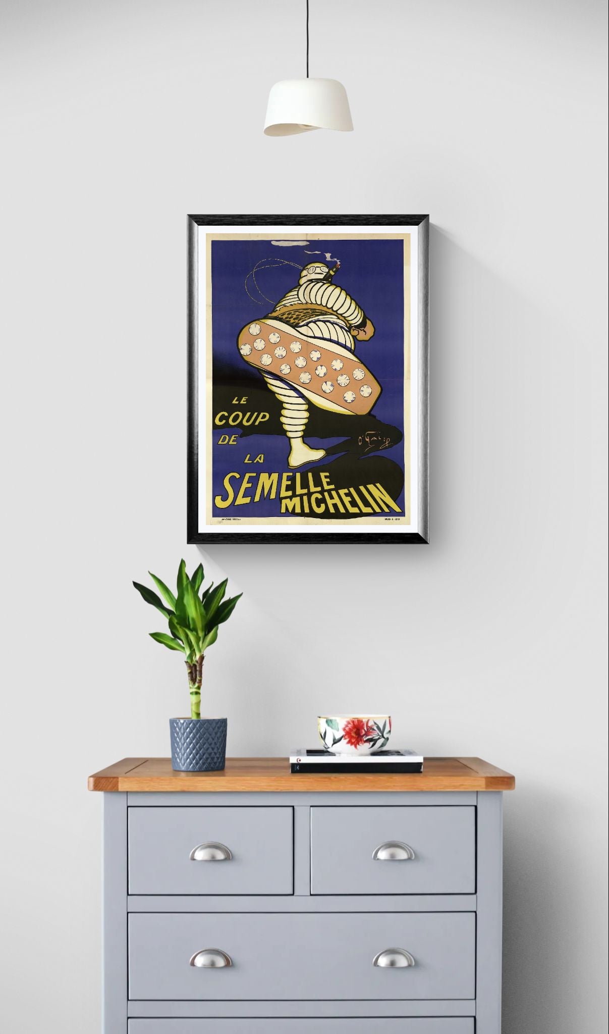 Framed artwork above a wooden dresser with a plant and small table on a light gray wall.