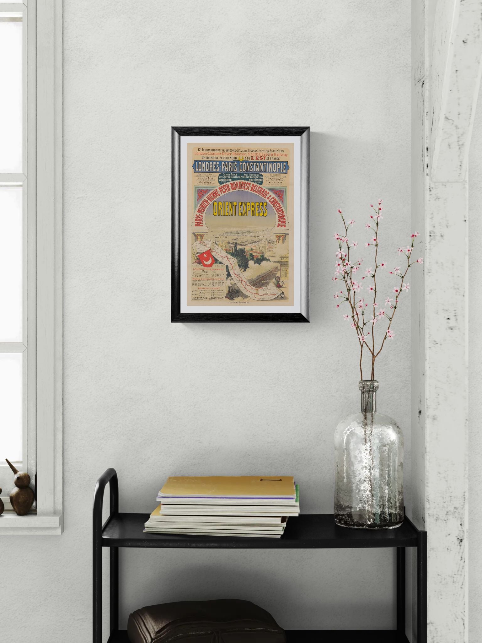 Framed vintage poster on a wall above a black shelf with books and a vase.