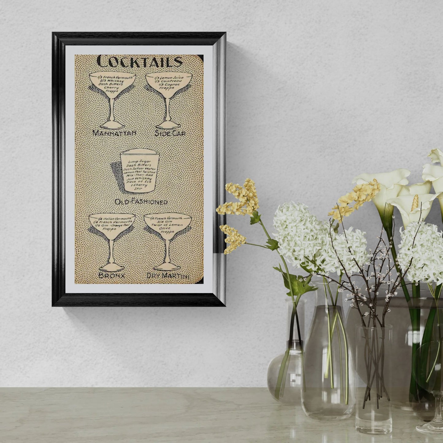 Framed cocktail recipe print on a wall with decorative vases and flowers.