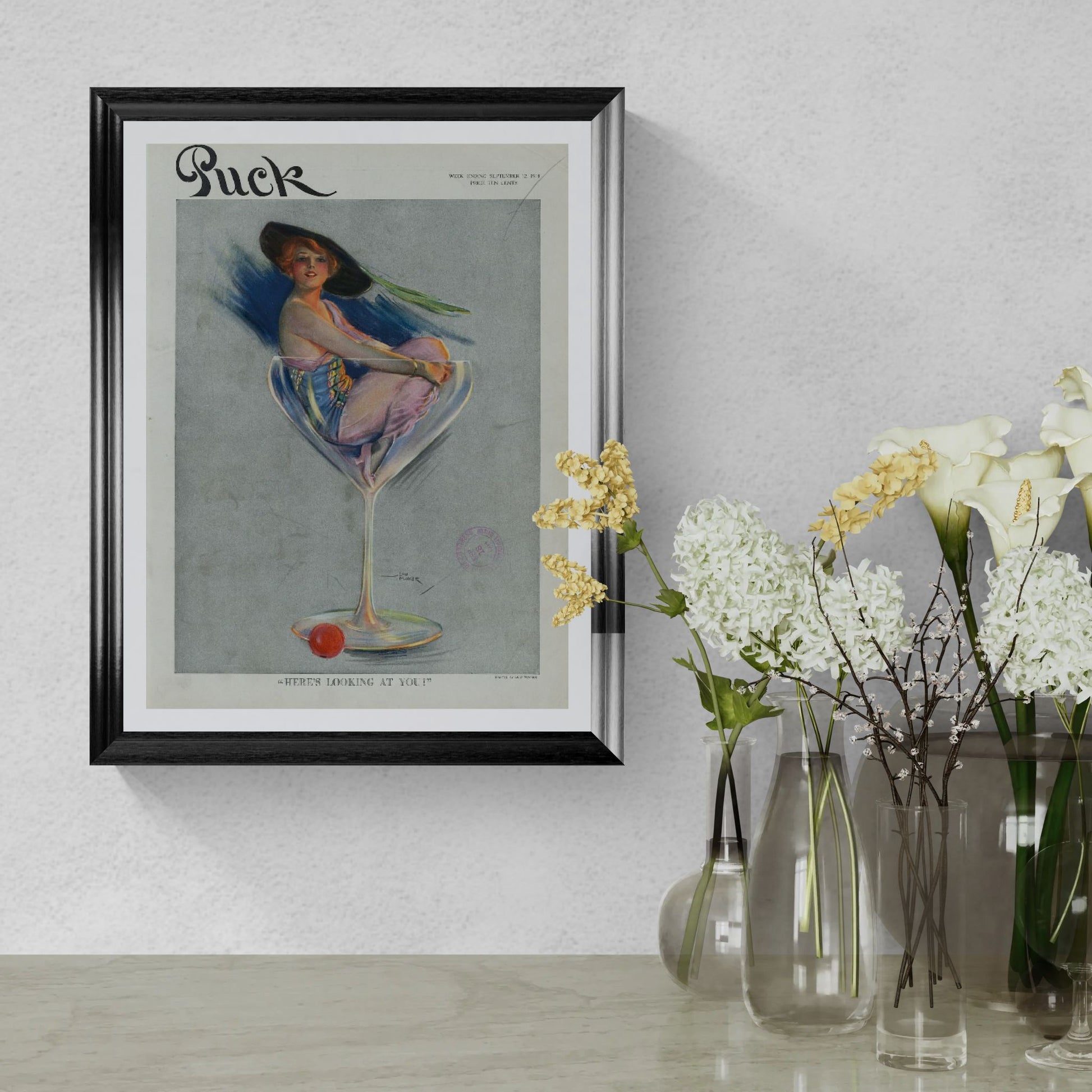 Framed vintage magazine cover of 'Puck' with decorative vases and flowers on a light gray wall.
