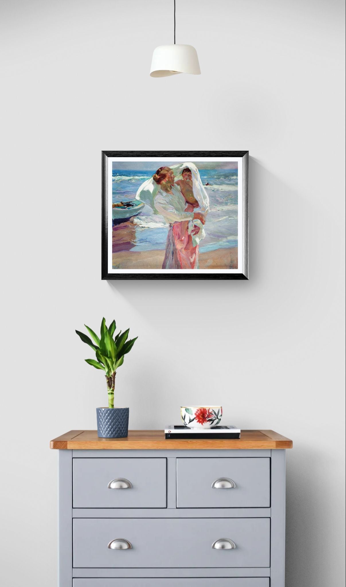 Framed artwork on a wall above a wooden dresser with a plant and small table.