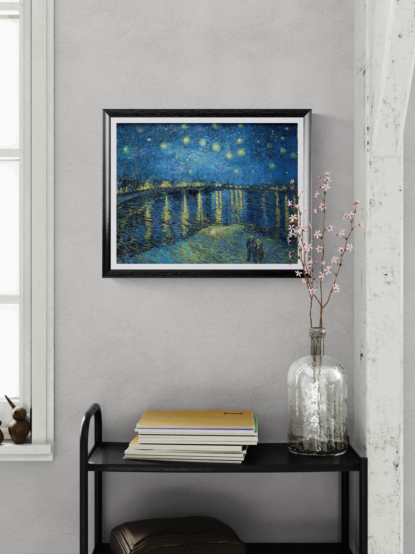 Framed artwork of a starry night scene on a wall above a black shelf with books and a vase.