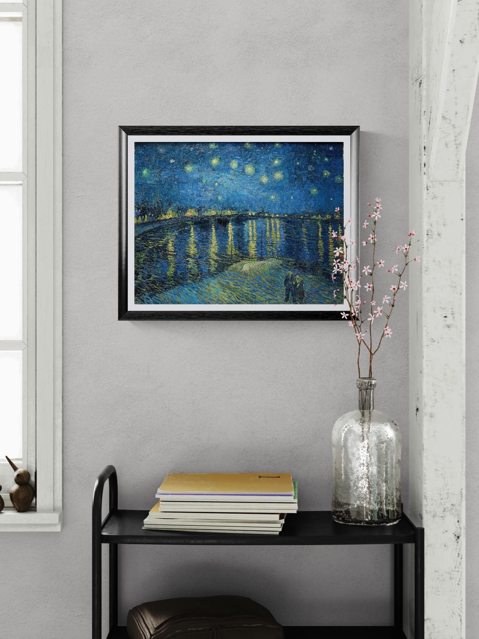 Framed artwork of a starry night scene on a wall above a black shelf with books and a vase.