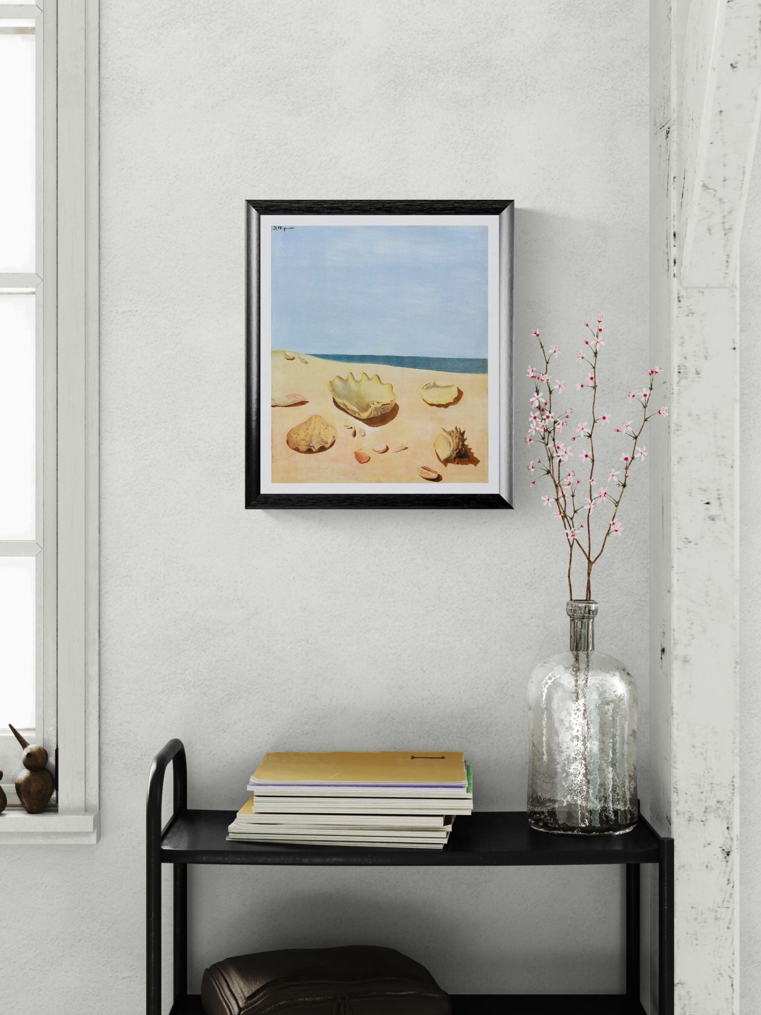 Framed beach scene print on a wall above a black shelf with books and a vase.
