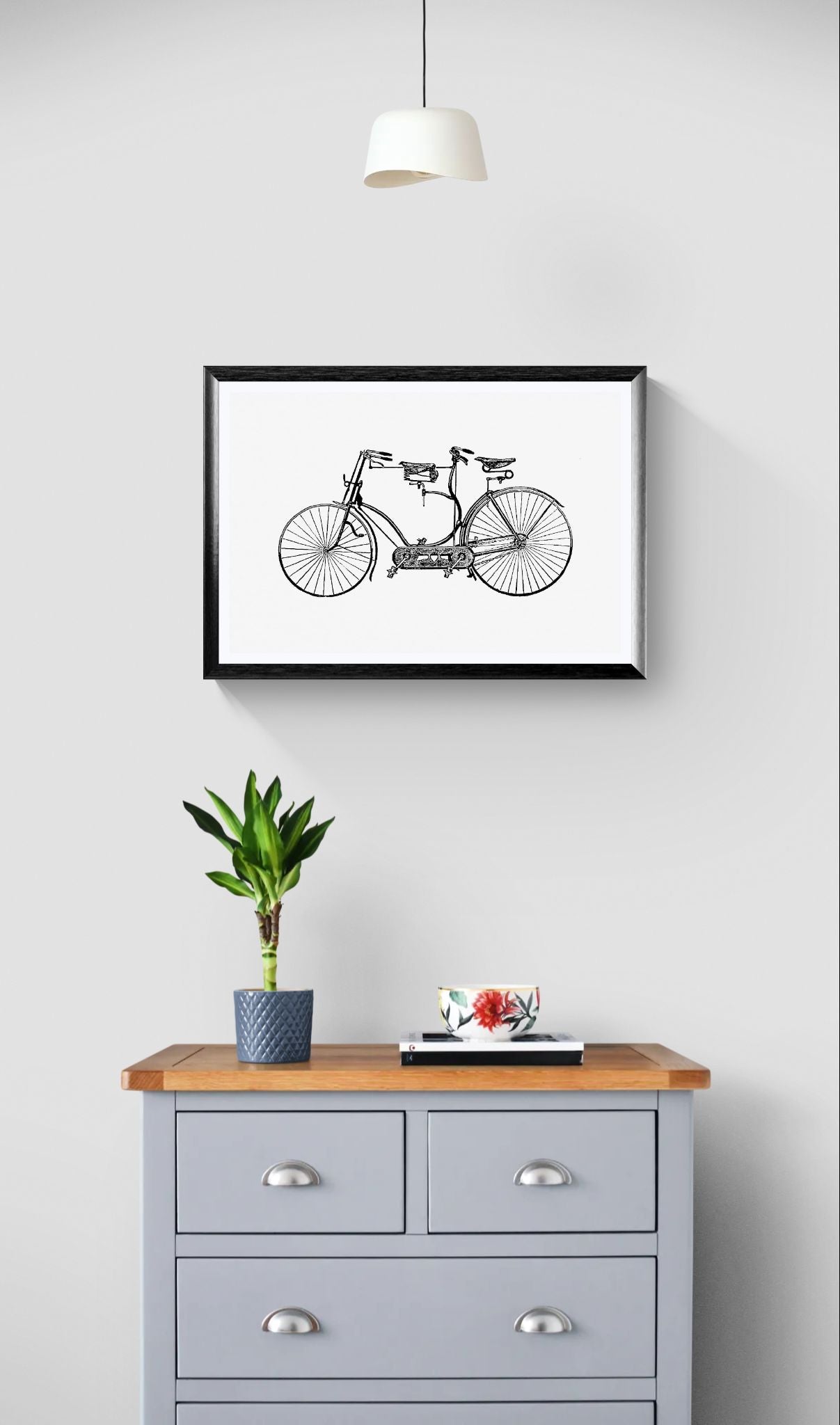 Framed black and white bicycle print above a wooden dresser with a plant and small table on top.