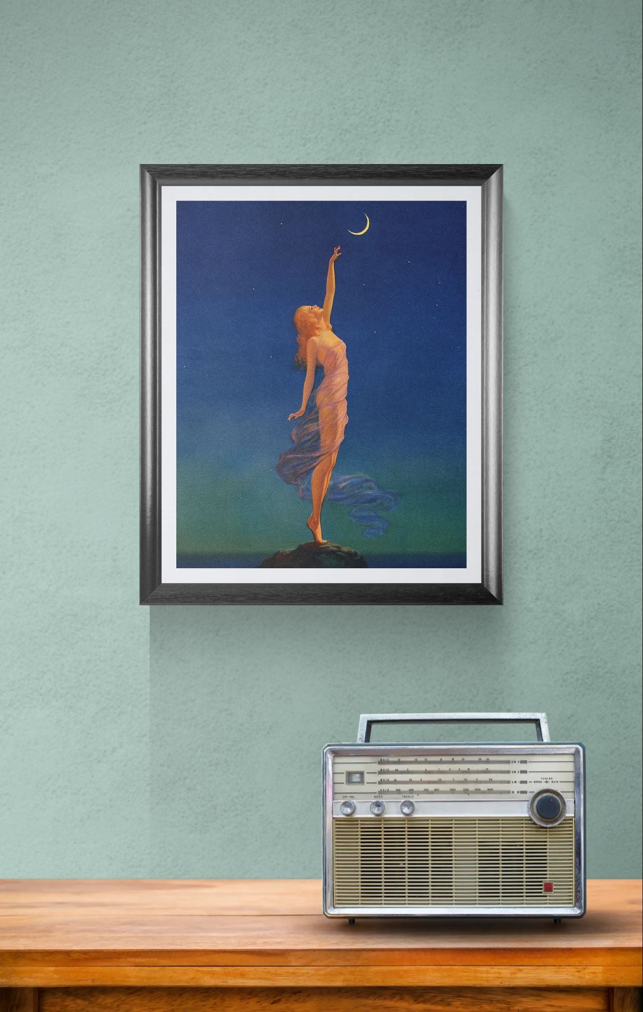 Framed artwork of a woman reaching for the moon above a vintage radio on a wooden surface.