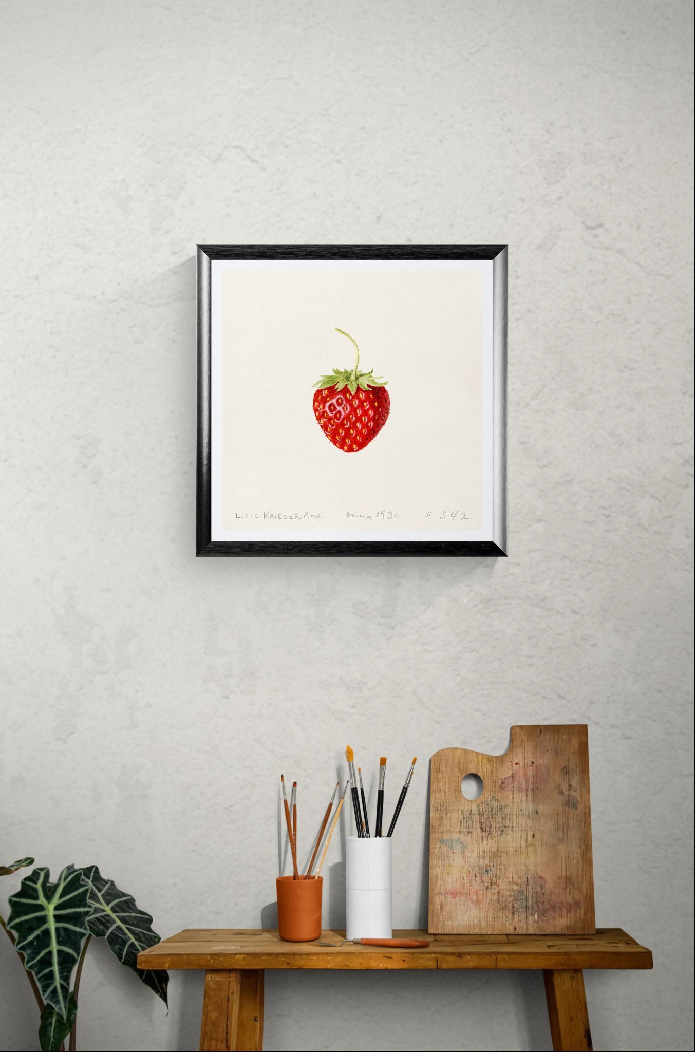 Framed artwork of a strawberry on a wall above a wooden table with art supplies.