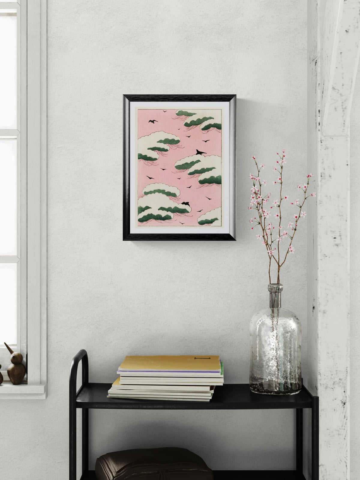 Framed artwork on a wall above a black shelf with books and a vase.