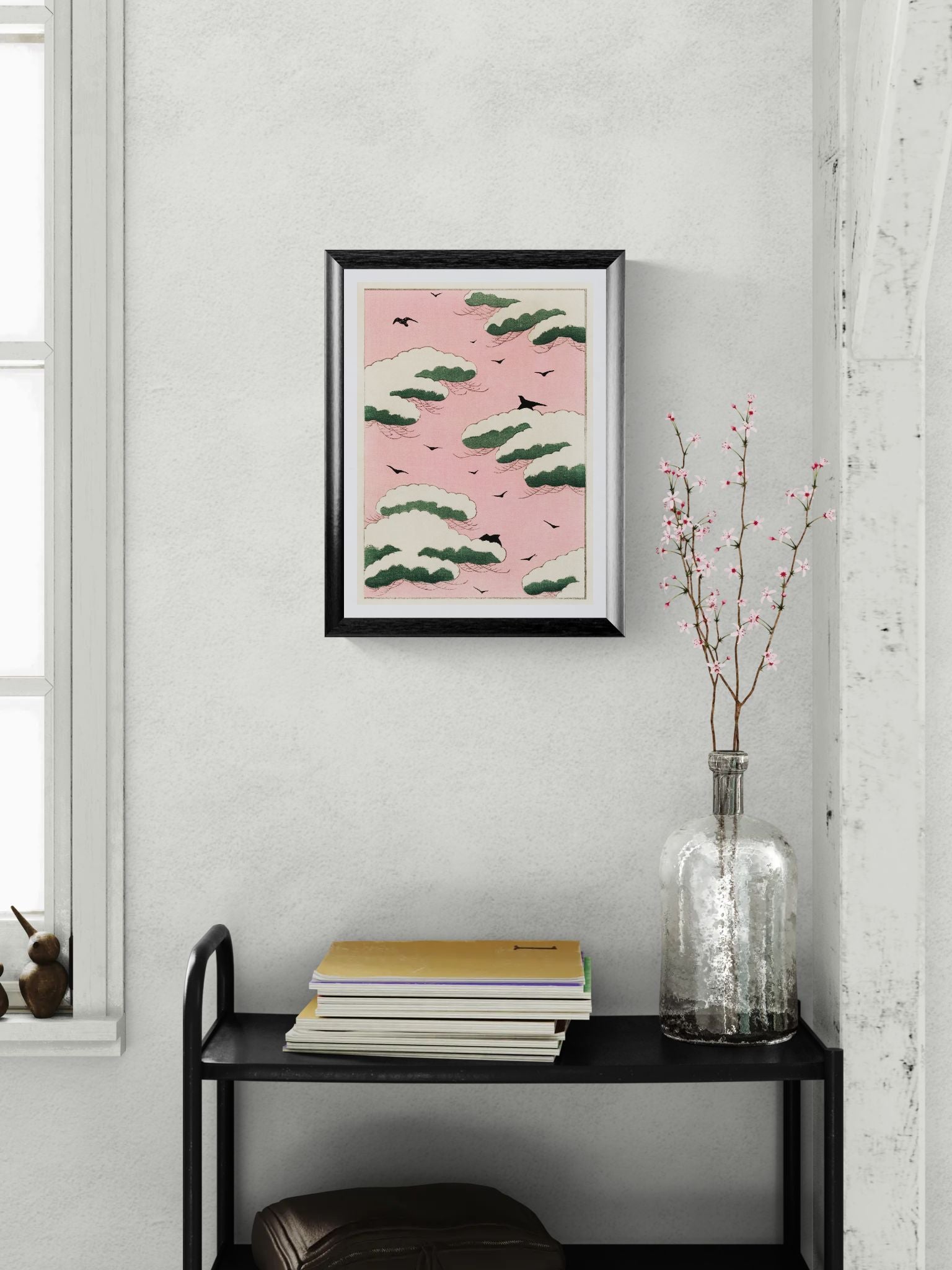 Framed artwork on a wall above a black shelf with books and a vase.