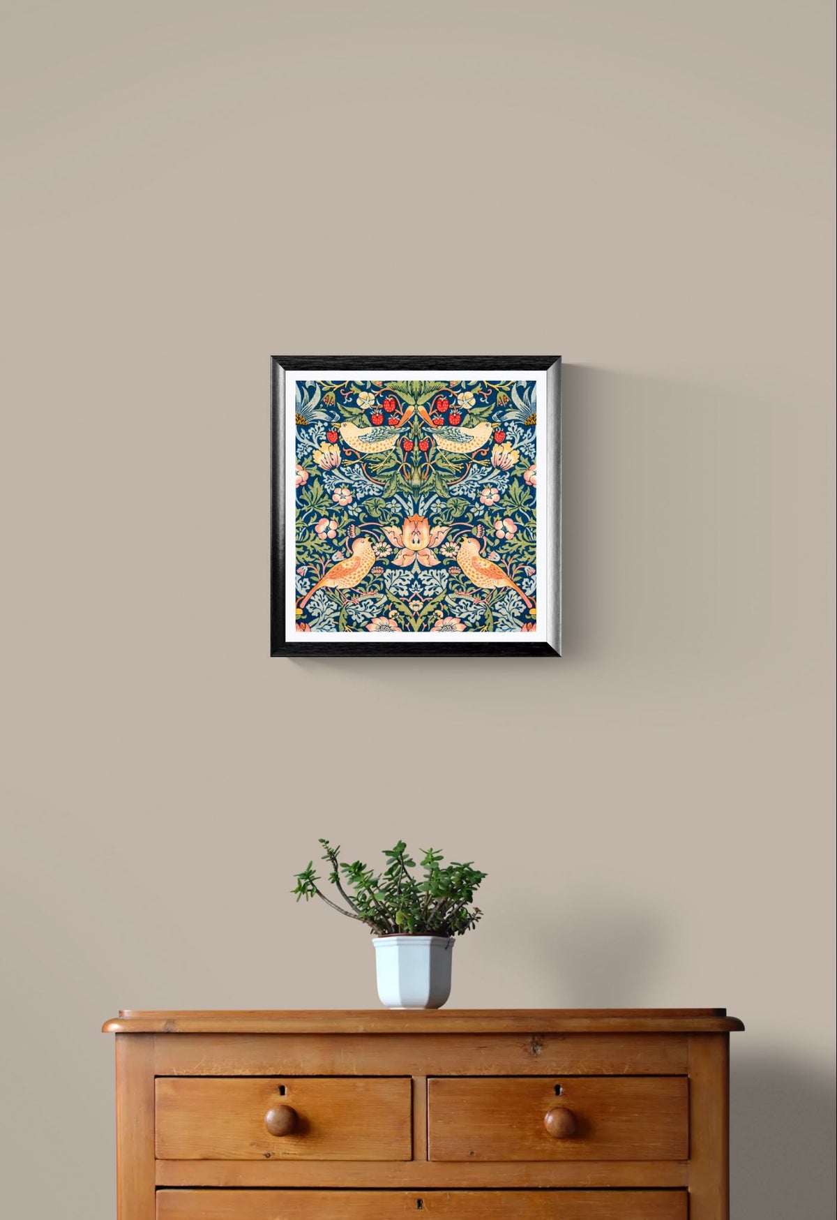 Framed artwork with floral and bird design on a beige wall above a wooden dresser with a plant.